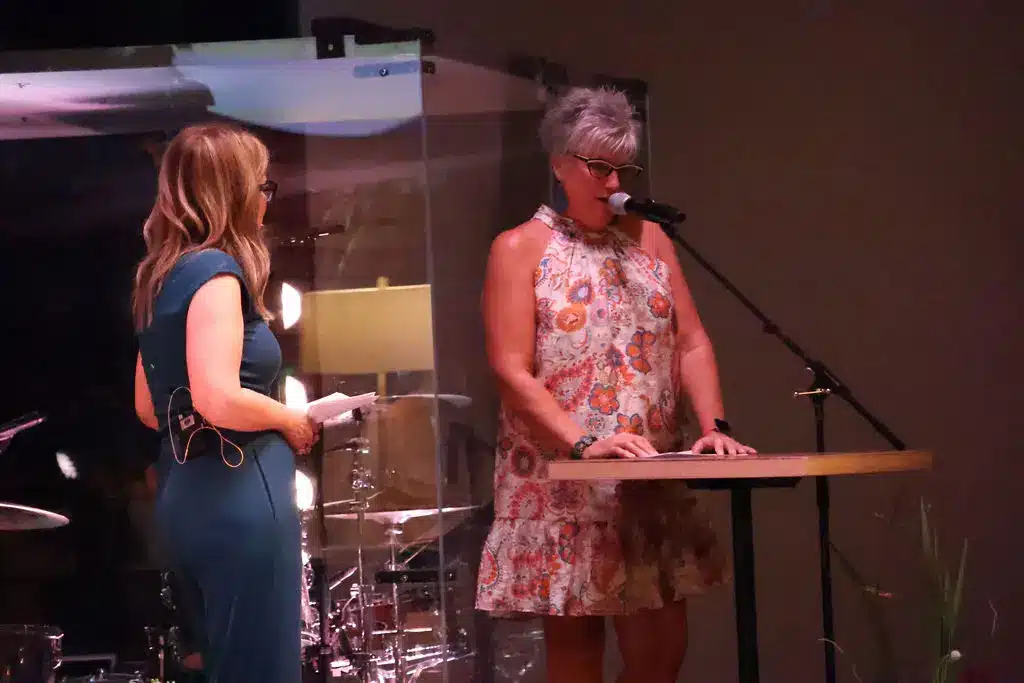 Event speaker addressing the audience from a podium while another presenter listens on stage during a community gathering