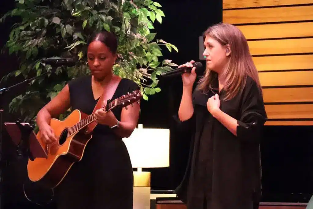 Two women performing on stage, one playing acoustic guitar and the other singing into a microphone during a live event