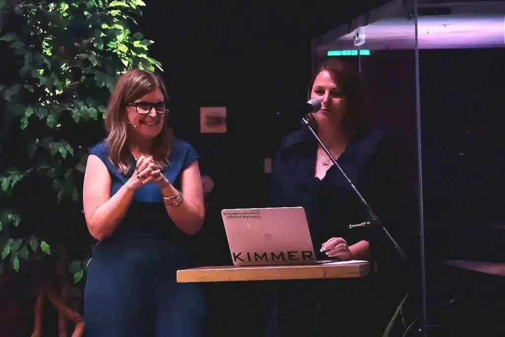 Two women standing on stage during a presentation, one speaking at a microphone beside a laptop, at a formal evening event